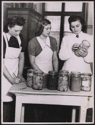Die Leiterin der Küche mit einem Abzeichen des Deutschen Frauenhilfsdiensts und eine zwei angehende Hausgehilfinnen bereiten eingewecktes Obst vor