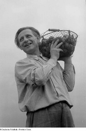 Porträt eines lachenden Mannes (Parteisekretär des Kaufhauses des Friedens) mit einem Kartoffelkorb auf der Schulter