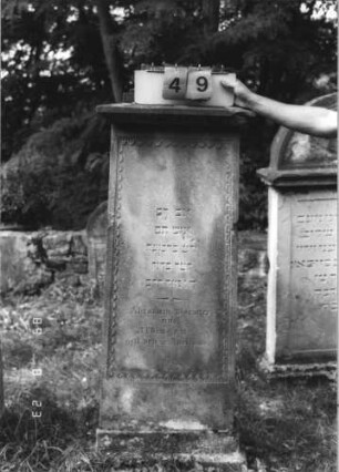 Grabstein 49 (Index: 100110864) Material: Sandstein Zustand: Giebel leicht beschädigt, leicht verwittert Maße HxBxT: 157x58x54 cm Beschreibung: Stele, Sockel getreppt, Giebel mit Aufsatz?, Schriftfeld eingetieft mit umlaufendem Dekor: Grabstein 49