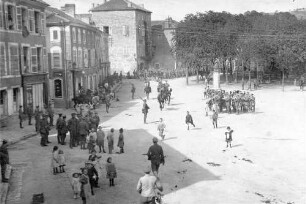 Ein auf einem größeren Platz spielendes Musikkorps; im Hintergrund marschieren Soldaten ein