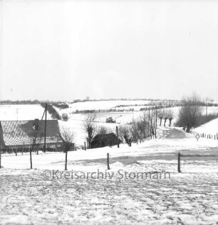 Steensrade: Gemeindeweg in Richtung Meddewade: im Vordergrund Wiese und Zaun: links Häuser: im Hintergrund Bauarbeiten mit Bagger an Durchbruch: Winter