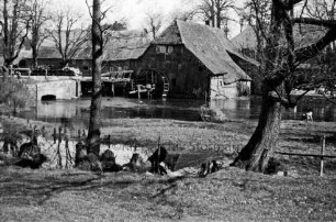 Grander Mühle mit Nebengebäuden: verfallenes Gebäude und beschädigtes Mühlrad: links Brücke: davor Mühlenteich und Bäume: rechts Kind am Wasser