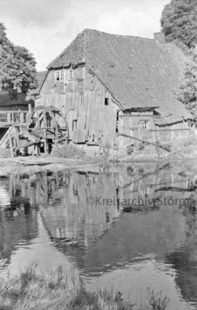 Grander Mühle mit Nebengebäuden: verfallenenes Gebäude und beschädigtes Mühlrad: links Holzbrücke: vorn Mühlenteich: hinten Bäume