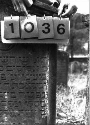 Grabstein 1036 (Index: 100098976) Maße HxBxT: 119x60x12 cm Symbolik: Trauerkranz Besonderheit: kulturhistorisch wertvoll / hist.-wiss. Gründe (Gemeindestellung): Grabstein 1036 (Nachholaufnahme)