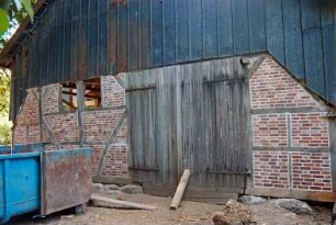 Dorfplatz (2): Fachwerkscheune: Sanierung: Giebelseite, Teilansicht: Schwellbalken auf Feldsteinfundament, Gefache mit neuer Ausmauerung, Scheunentor, oben Blechverkleidung: davor Schuttcontainer: 8. August 1994