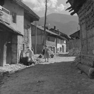Alltagsbild. Frauen unterwegs in Bansko. Gestelle für das Trocknen der Tabakblätter am Wohnhaus und ein öffentlicher Brunnen in Bansko
