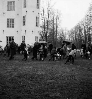Schlosspark: Ostereiersuchen: Kinder suchen im Regen: Erwachsene mit Regenschirmen: links Schloss