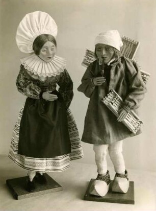 Zwei Spielfiguren in Stoffkleidung. Paar in Volkstracht. Frau mit Kleid, Schürze und weiter Haube. Mann mit Pfeife, Rock, Haube, Holzschuhen und Werkzeugen. Halbtotale