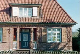 Kamp 19: traufständiges Wohnhaus aus Backstein: Straßenfront, Teilansicht: Eingangsloggia mit Tür, darüber Flachgaube, Sprossenfenster mit Fensterläden: vorne Jägerzaun, Gartenpforte