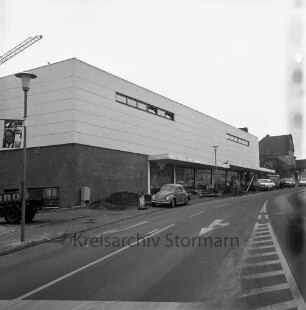 Käselau: Kaufhaus: Bahnhofstraße: vor Fertigstellung: Außenansicht: Straßenfront: Arbeiter beim Herrichten von Fußgängerweg und Parkbuchten: Straßenlampen, Fahrbahnmarkierungen, links Wahlplakat