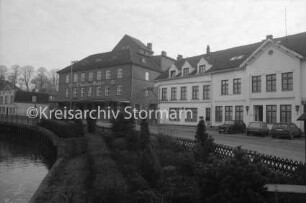 Handel: Claudius-Mühle: Matthias-Claudius-Straße: links Produktionsgebäude mit Laderampe, rechts Verwaltungsgebäude: davor rechts drei parkende Pkws: vorn Grünanlage und Jägerzaun am Herrenteich: hinten links Gebäude: 12. Dezember 1985