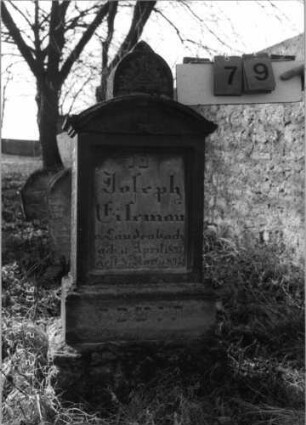 Grabstein 79 (Index: 100116224) Material: Sandstein Zustand: gut Maße HxBxT: 100x35x21 cm Beschreibung: aufgesockelte Stele mit eingezogenem Segmentbogenabschluß und Aufsatz, eingetieftes Schriftfeld Besonderheit: Kindergrab: Grabstein 79