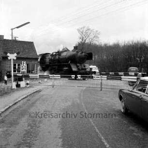 Bahnübergang Lübecker Straße bei Forst Kneeden: Bahnstrecke zwischen Hamburg und Lübeck: Schrankenposten 54: Schranken geschlossen: Durchfahrt einer Dampflokomotive: wartende Autos: Andreaskreuze, Schrankenwärterhäuschen, Lichtmast: hinten Bäume