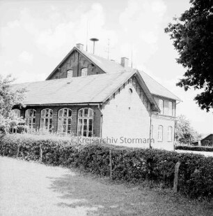VVolksschule: Außenansicht der Gebäude: vorn Hecke und Zaun: rechts oben Baumäste