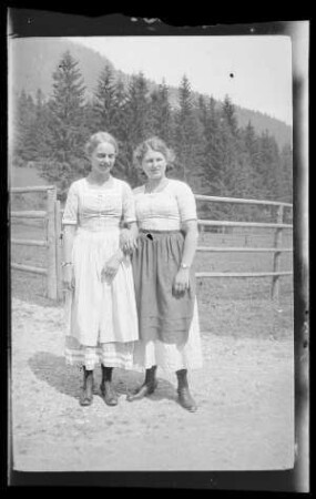 Gegend von Schliersee (Lkr. Miesbach): 2 sich einhakende junge Frauen im hellen Dirndl auf einem Weg vor einem Viehgatter