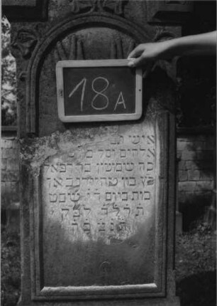 Grabstein 18 (Index: 99801905) Material: Sandstein Zustand: gebrochen und repariert mit Textverlust Maße HxBxT: 190x50x20 cm Beschreibung: Stele, getreppter Sockel, Rundbogen, eingetieftes Schriftfeld mit Rundbogen Symbolik: segnende Hände, Blätterfries: Grabstein 18A