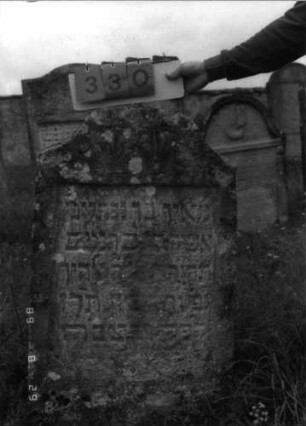Grabstein 330 (Index: 100107760) Material: Kalkstein Zustand: stark verwittert, teilweise Textverlust Maße HxBxT: 112x63x18 cm Beschreibung: Stele, geschwungen eingezogener, oben abgeplatteter Korbbogenabschluß, Schriftfeld rechteckig eingetieft Symbolik: segnende Hände: Grabstein 330