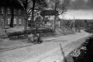 Groß Barnitz: Dorfstraße mit Kopfsteinpflaster: links zwei Frauen mit Fahrrädern: dahinter Hecke, Vorgärten mit Bäumen und Bauerngehöfte