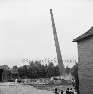 Stadtwerke Bad Oldesloe: Gaswerk: Lübecker Straße: Sprengung des Schornsteins des abgerissenen Kraftwerks: rechts Gebäudeecke und Köpfe von Zuschauern: dahinter geneigter Schornstein mit Gastank, am Fuß Rauchwolken: hinten Bauschutt: links teilweise abgerissenes Gebäude: hinten Bäume: 10. Juni 1971