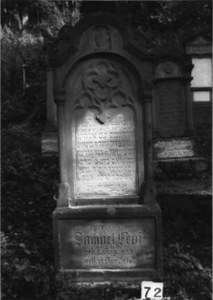 Grabstein 72 (Index: 100079953) Material: Sandstein Zustand: leicht verwittert mit Textverlust Maße HxBxT: 159x60x23 cm Beschreibung: ädikulaartige Stele mit Halbsäulen, Sockel, eingezogener Rundbogenabschluß mit Eckakroteren, Schriftfeld eingetieft Symbolik: Levitenkanne, Ranken, Maßwerk: Grabstein 72