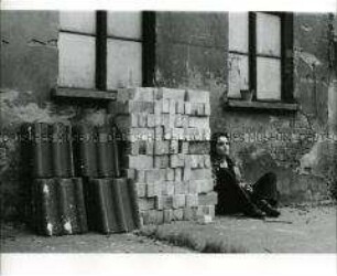 Jugendliche sitzt neben aufgestapelten Mauersteinen an eine Hausfassade gelehnt (Altersgruppe 18-21)