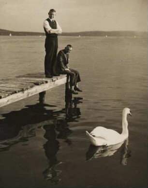 An einem See. Zwei Männer befinden sich auf einem Bootssteg. Sie betrachten einen Schwan