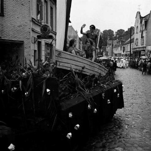 Karpfenfest: Umzug: Paul-von-Schoenaich-Straße: Festwagen, drei Frauen in Fischerboot, Schilf: hinten Fußgruppe Köche: rechts am Straßenrand Zuschauer, mit Regenschirmen: am linken Straßenrand Schuhgeschäft Wilke: im Hintergrund Fahnenschmuck, Wimpelgirlanden, Bäume des Kirchhofs, 8. Oktober 1961