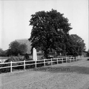 Dorfring: links Ehrenmal 1939-1945 mit Grünanlage: davor Zaun: rechts Friedenseiche: hinten an Straße Häuser mit Gärten: hinten rechts Milchkannen