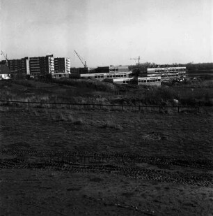 Schule am Masurenweg [Dörfergemeinschaftsschule, Grund- und Hauptschule]: Rohbau: links Haus und Wohnblocks: dahinter Baukräne: vorn unbefestigtes Gelände, Zäune, Garten, Erdhaufen und Wiese: 11.11.1972