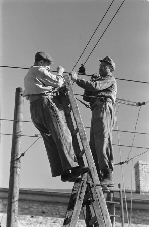 Energieversorgung. Arbeiten an einer Oberleitung. Ansicht mit zwei Arbeitern, von einer Leiter aus an den Leitungen arbeitend