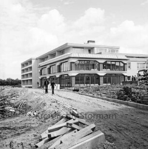 Seniorenwohnanlage Rosenhof I: Hoisdorfer Landstraße: Gebäudekomplex vor Fertigstellung: in der Mitte Hauptgebäude mit Gemeinschaftseinrichtungen: dahinter Appartmenthäuser: vorn Baumaterial, unbefestigter Weg: darauf zwei Personen: rechts Baumaschinen
