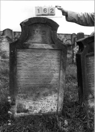 Grabstein 162 (Index: 100107436) Material: gelber Sandstein Zustand: stark verwittert und versalzen, weitgehender Textverlust Maße HxBxT: 132x71x15 cm Beschreibung: Stele in Sonderform, Schriftfeld eingetieft: Grabstein 162