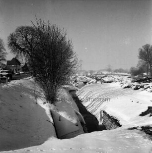 Mühlenbach: stillgelegter Ausbau: rechts Erdhaufen: links an Böschung Bäume: hinten links Stormarnstraße mit Lieferwagen und Pkw: dahinter Tankstelle: hinten rechts eingezäunte Weiden: Winter