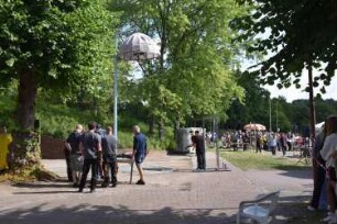 Bad Oldesloe: Kindervogelschießen: Exer: An Mast aufgehängter Vogel für die männlichen Teilnehmer in Geschossfang: davor Aufsichtspersonen beim Spannen der Armbrüste: mittig Teilnehmer, im Hintergrund Schießstand der Mädchen, rechts Zuschauer: 13. Juli 2023