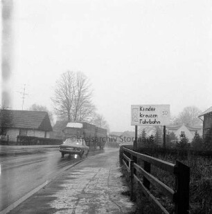 Hauptstraße: rechts Zaun. dahinter in Vorgarten Hinweisschild auf Kinder: hinten Mitte Lkw und Pkw: dahinter parkender Lkw: hinten rechts und links Wohn- und Geschäftshäuser