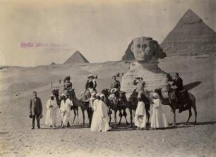 Gizeh, Ägypten. Touristen auf Kamelen posieren mit einheimischen Führern vor der Sphinx und den Pyramiden von Gizeh