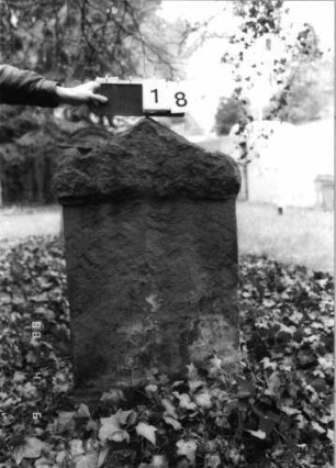 Grabstein 18 (Index: 99793026) Material: Sandstein Zustand: sehr stark verwittert mit fast vollständigem Textverlust Maße HxBxT: 111x64x12 cm Beschreibung: Stele, eingezogener, giebelartiger Abschluß mit Eckakroteren: Grabstein 18