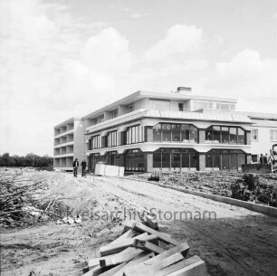 Seniorenwohnanlage Rosenhof I: Hoisdorfer Landstraße: Gebäudekomplex vor Fertigstellung: in der Mitte Hauptgebäude mit Gemeinschaftseinrichtungen: dahinter Appartmenthäuser: vorn Baumaterial, unbefestigter Weg: darauf zwei Personen: rechts Baumaschinen