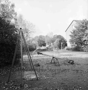 Kinderspielplatz: Wippe, Schaukel, Klettergerüst, Hecke: hinten Jägerzaun, Wohnhaus, parkende Autos