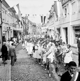 Kindervogelschießen: Umzug: Hindenburgstraße: Jungen mit Schärpen, Mädchen mit Blumenbögen, hinten Spielmannszug der Stadtschule: an den Straßenrändern Zuschauer: über Straße Wimpelgirlanden, an Fassaden Fahnenschmuck: an linker Straßenseite PFAFF-Nähmaschinengeschäft, Commerzbank, Friseursalon Lübcke, hinten Gaststätte "Bierquelle": an rechter Straßenseite Sattlerei Polsterei: im Hintergrund Hagenstraße, 2. Juli 1963