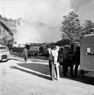 Brand: Gutshof Altfresenburg: Bullenstall mit Stroh- und Heuboden: vorn rechts Feuerwehrmänner an Löschfahrzeug beim Entladen von Wasserschläuchen und Wasserpumpe: hinten links Feuerwehrmann: dahinter weitere Feuerwehrfahrzeuge: hinten links brennendes Gebäude in Rauchschwaden: davor weitere Feuerwehrleute: hinten rechts Bäume