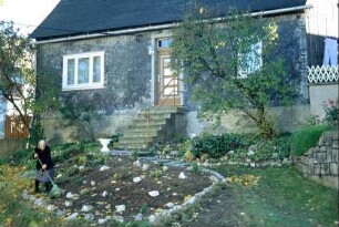 kleines Haus, davor Treppe, Fliederbusch; Frau im Garten