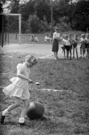 Kinder bei einem Wettbewerb am Kindertag, Dessau : Ein Mädchen beim Eierlauf auf einem Sportplatz, im Hintergrund stehen weitere Kinder und eine Frau
