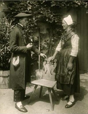 Ehepaar stehend vor historischem Holzstuhl, Frau rechts gestützt, Mann mit Pfeife links daneben. Halbtotale