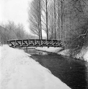 Beste: hinten Holzbrücke für Fußgänger: rechts Kurpark mit Bäumen: links Baumreihe: dahinter Gebäude: Winter