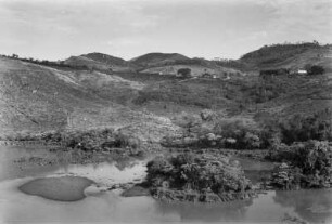 Flusslandschaft mit Hügeln : Im Vordergrund ein Flusslauf mit baumbewachsenen Inseln. Im Hintergrund Grasflächen und Bäume auf den Hängen von Hügel, sowie Gebäude.