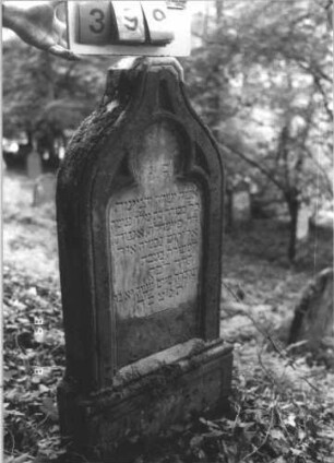 Grabstein 399 (Index: 99799632) Material: Sandstein Zustand: verwittert, Farbreste in den Lettern, Mittelaufsatz fehlt Maße HxBxT: 122x46x14 cm Beschreibung: Stele, Abschluß Eselsrücken, Mittelaufsatz fehlt, Schriftfeld eingetieft in Maßwerkform, Sockel: Grabstein 399