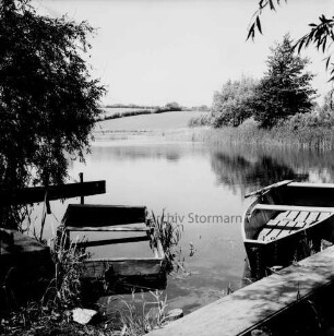 Moorteich: im Vordergrund Steg mit zwei Kähnen: links Busch: hinten Bäume und Knicklandschaft