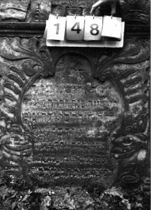 Grabstein 148 (Index: 99751968) Material: Sandstein Zustand: stark verwittert mit Textverlust Maße HxBxT: 142x99x10 cm Beschreibung: Stele, in Voluten auslaufend. Rundbogenaufsatz, Schriftfeld in Wappenschildform Symbolik: drei Kronen: Grabstein 148B