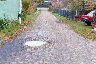 Fritz-Berodt-Straße: Fahrbahn mit Pflasterung: beiderseits Gärten, vorne links Gebäude, hinten Verkehrsschild, Einmündung in Straße